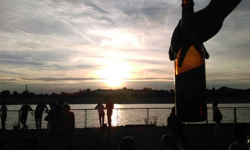 vista do rio reno a partir de uma escadaria em Dusseldorf, alemanha. COntra o sol está uma mão segurando uma cerveja