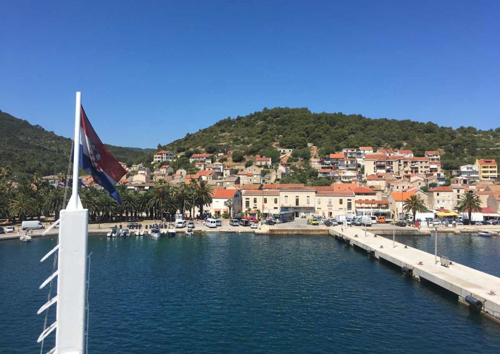 Ferry boat atracando em Vis. Bandeira da Croácia em primeiro plano e o centro da ilha em segundo plano, com prédios bege e montanha ao fundo